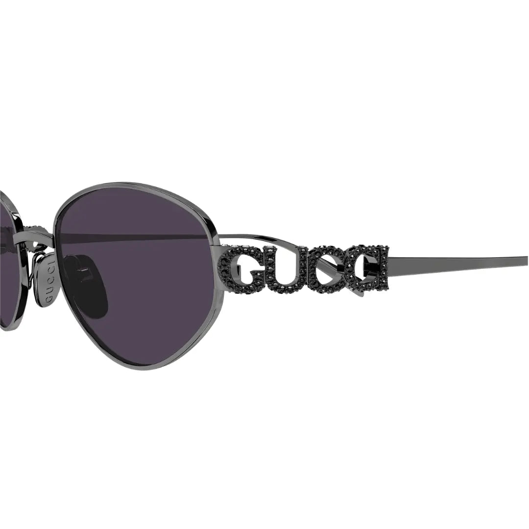 Close-up side view of Gucci sunglasses with dark gray metal frames, dark lenses, and a prominent rhinestone-embellished GUCCI logo on the temple arm, shown against a plain white background.